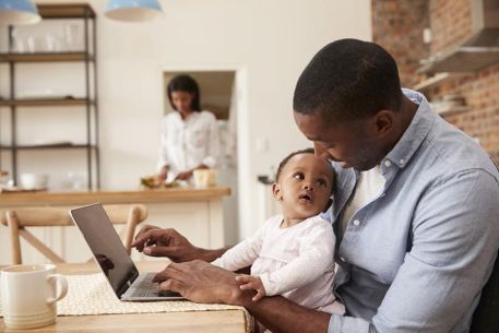 A man is setting at the table at home studying on his laptop with his little child on his lap looking up at him.